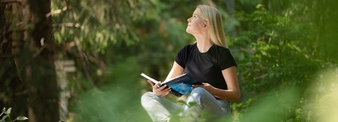 Nuori ihminen lukee kirjaa metsässä ja katsoo kohti taivasta.