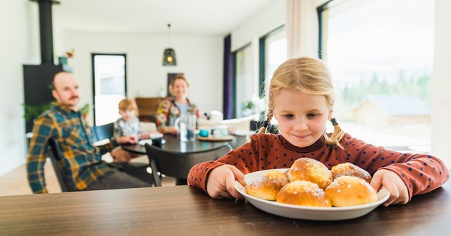 Tyttö pitää kiinni pullalautasesta, joka on pöydällä ja katsoo pullia. Äiti, pikkusisko ja isä istuvat taustalla ruokapöydän ääressä.