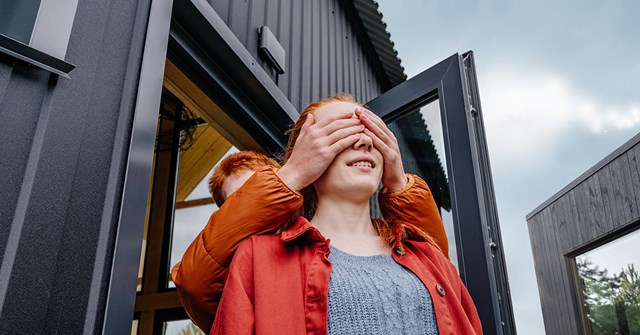 Two people stand in front of the house and the one behind covers the other's eyes with his hands.