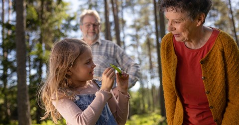 Lapsi ja isovanhemmat katsovat puusta tippunutta lehteä metsässä ja hymyilevät.