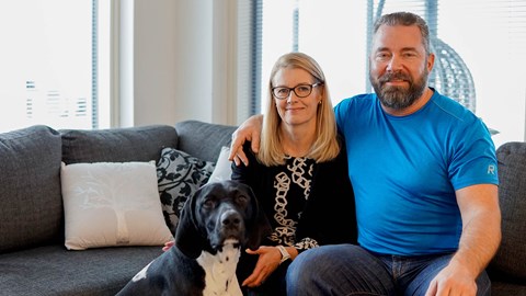 Minna and Pasi Alajoki are sitting on a gray sofa. In front of them sits the family's black and white dog.