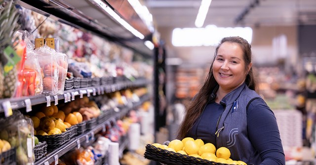 Nainen katsoo kameraan ruokakaupan kylmäosastolla. Hän hymyilee ja sylissä on hedelmäkori.